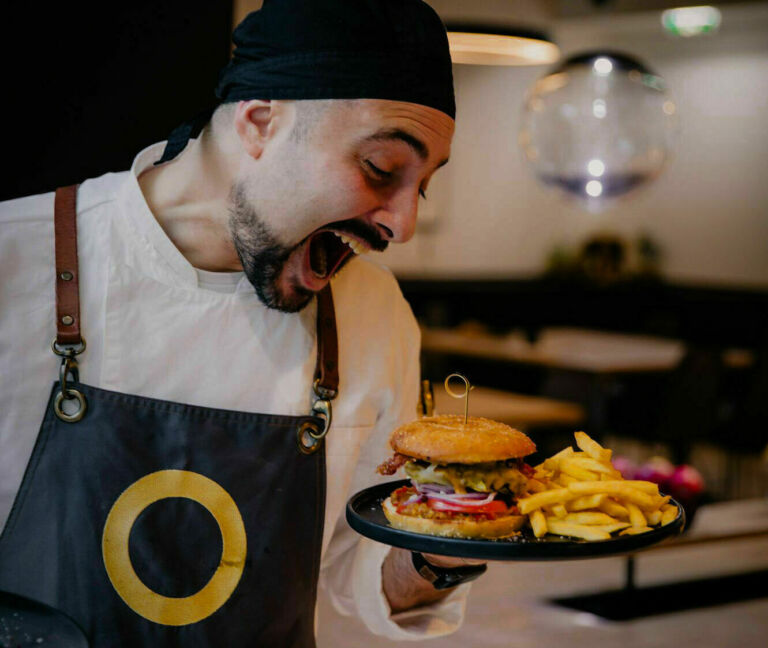 Koch in moderner Küche präsentiert begeistert einen Burger mit Pommes auf einem schwarzen Teller.