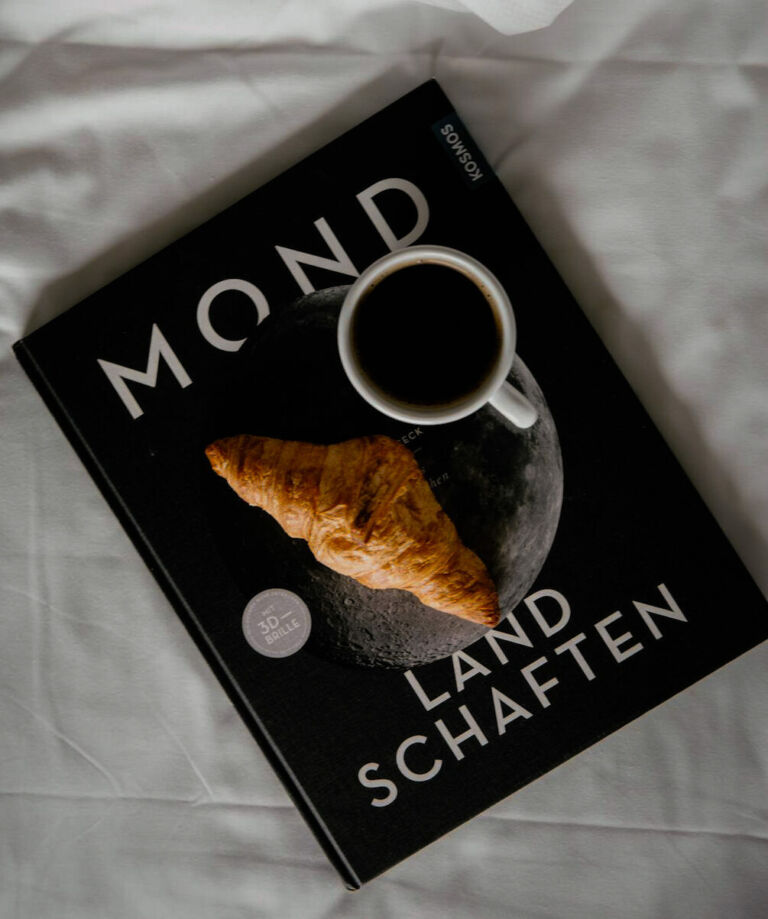 Croissant und Kaffee auf dem Buch "Mondlandschaften" auf weißer Bettwäsche.