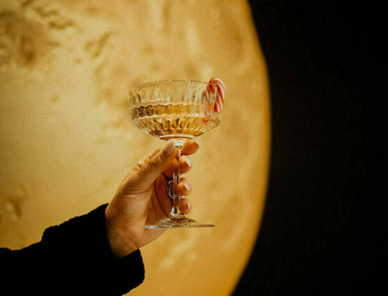 Hand hält ein Cocktailglas mit Zuckerstange vor mondähnlicher Kulisse.