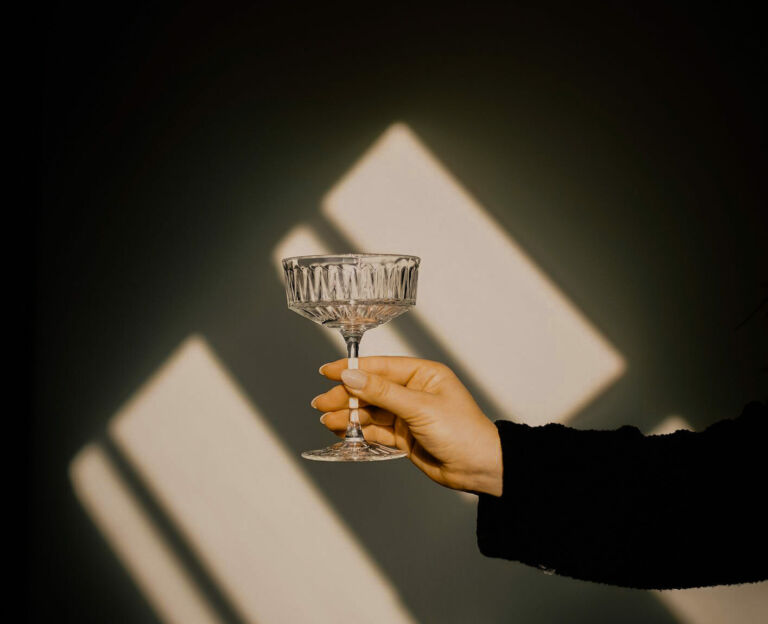 Hand hält ein leeres Cocktailglas vor einer Wand mit geometrischen Lichtmustern.